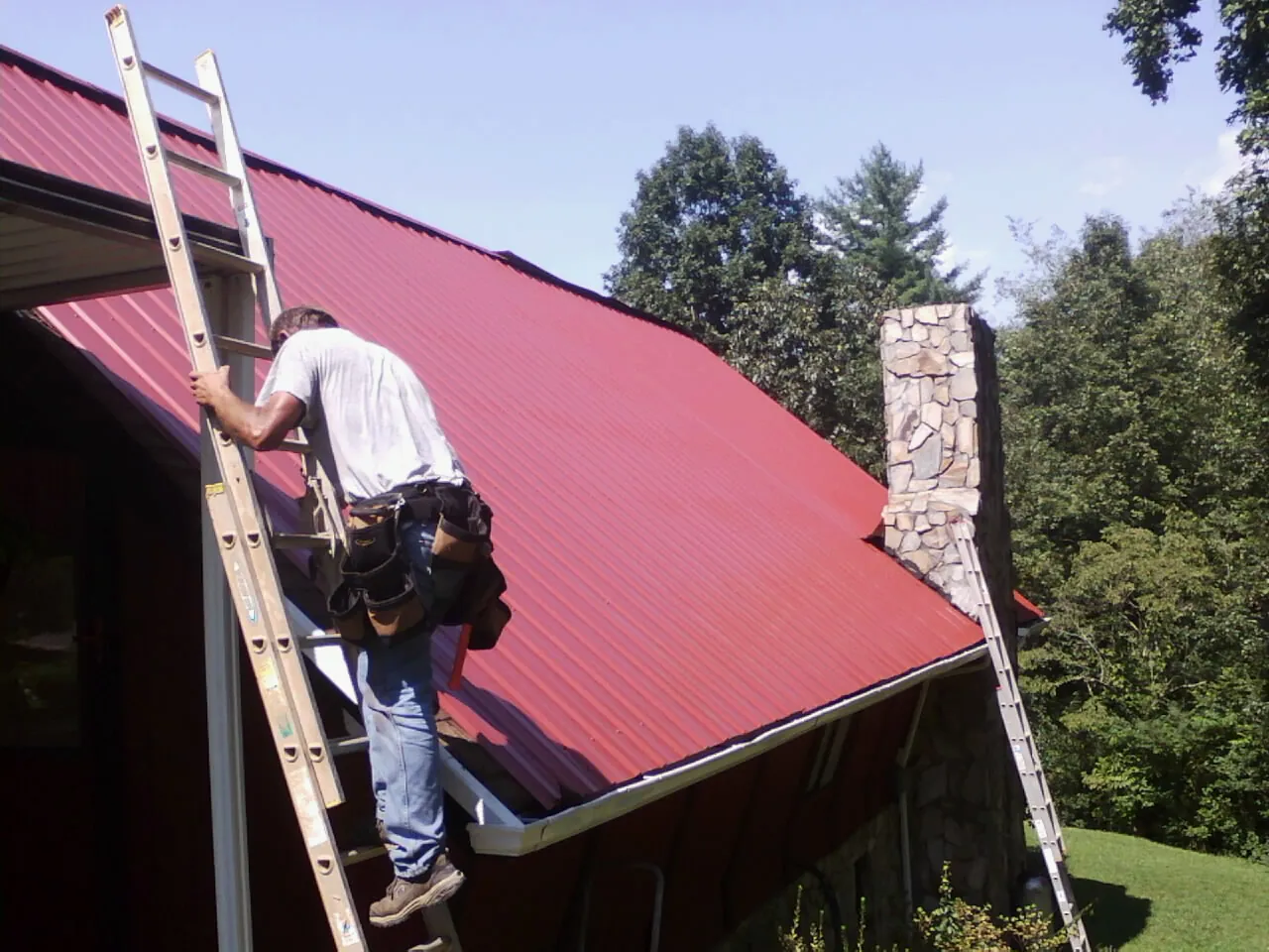 Roofer on a ladder beside a red metal roof and stone chimney — Soffit & Fascia Repair by Roofing Brothers in Harrietta